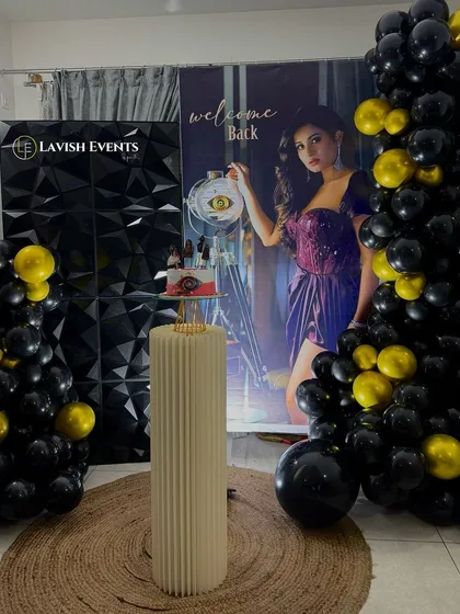 A full view of the welcome back party setup for a BBK contestant, featuring a 3D geometric wall, black and yellow balloons, and a personalized life-size banner.