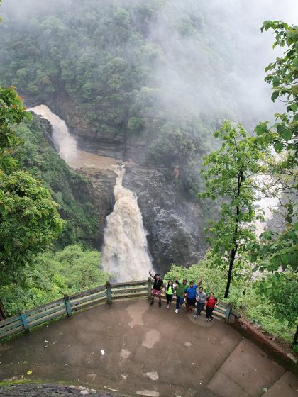 A group photo from a viewpoint overlooking a massive, muddy waterfall during the peak of monsoon. The scale of nature here is truly humbling.
