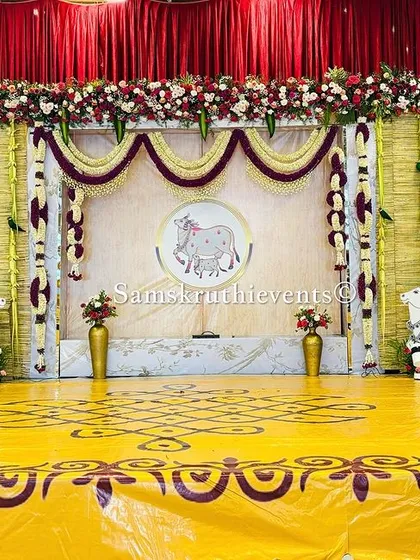 A grand stage with a Pichwai-inspired backdrop, featuring two large art panels and cow figures. The yellow floor with traditional rangoli patterns completes this magnificent setup.