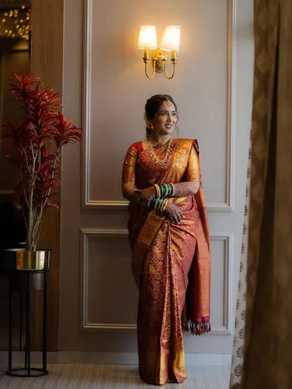 An elegant full-length portrait of the bride in her red and gold saree, standing by a beautifully decorated wall.