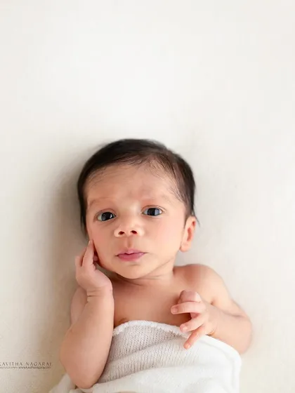 A two-week-old newborn looks up at the camera with a curious expression. The simple white wrap and background keep the focus entirely on the baby.