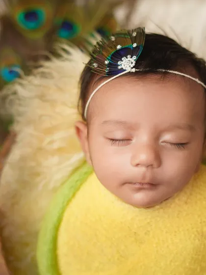 A close-up of a newborn dressed as little Krishna, with a peacock feather headband. This is a beautiful and traditional theme for a newborn photoshoot.