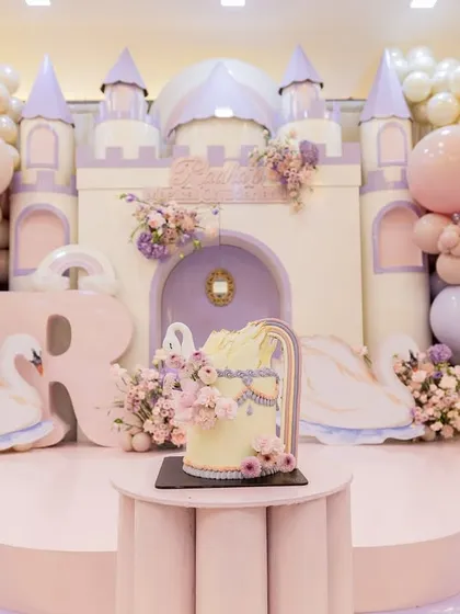 The main stage of the princess party, featuring a magnificent castle, whimsical swan figures, and a beautiful cake, all framed by pastel balloon clouds.
