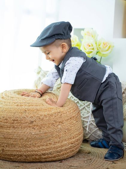 This little gentleman is having fun climbing on a jute pouf, dressed smartly in a vest and cap.