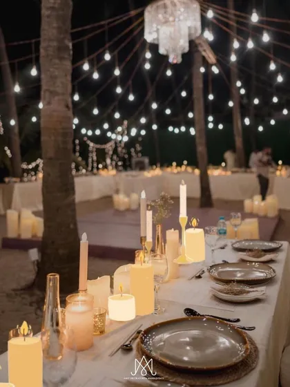 A close-up of a beautifully set table at the beach dinner, with rustic plates, candles of varying heights, and a canopy of string lights above.