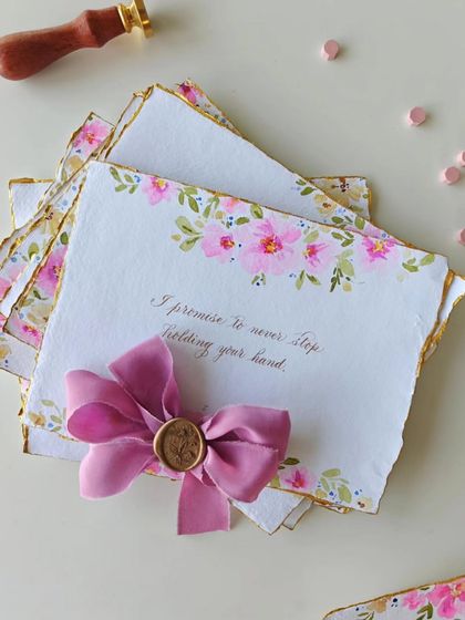 "I promise to never stop holding your hand." A single, powerful line from a set of handwritten vows, adorned with delicate pink watercolor florals and finished with a wax-sealed silk ribbon.