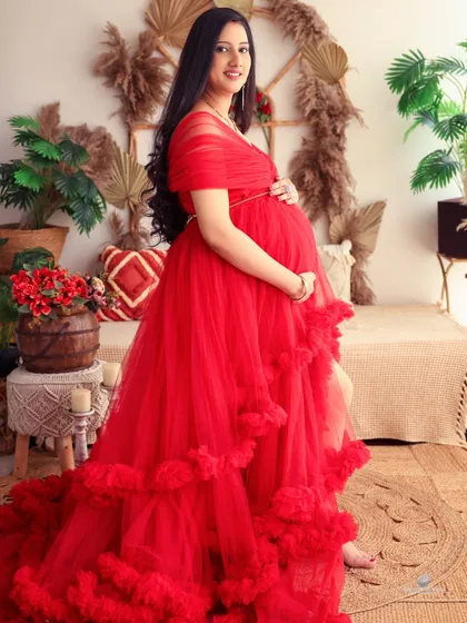 A classic standing portrait showcasing the full length of the vibrant red ruffled gown. The mom-to-be cradles her bump in our warm, boho-themed studio.