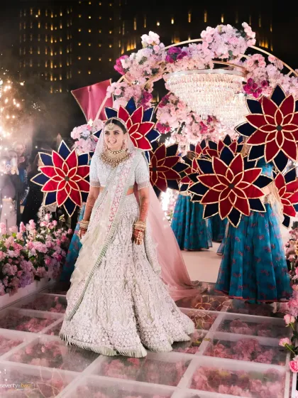 The bride's grand entrance at her Dubai wedding, walking down a reflective aisle under a canopy of flowers and a grand chandelier, surrounded by vibrant decor.