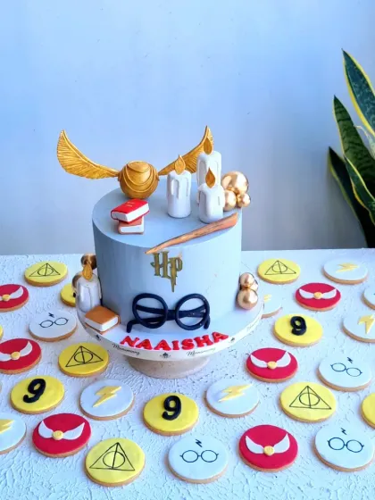 A magical Harry Potter themed birthday cake surrounded by custom-made cookies. The cake features iconic elements like the golden snitch, glasses, and a wand, while the cookies are decorated with symbols from the series, making it a perfect treat for any fan.