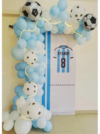 A simple and clean football-themed setup. The custom backdrop with an Argentinian jersey design is framed by a light blue and white balloon garland with fairy lights.