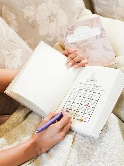 A lifestyle shot showing someone filling out a "Kindness Bingo" in their journal while relaxing on a couch. This image connects the product with positive, mindful activities and self-improvement.
