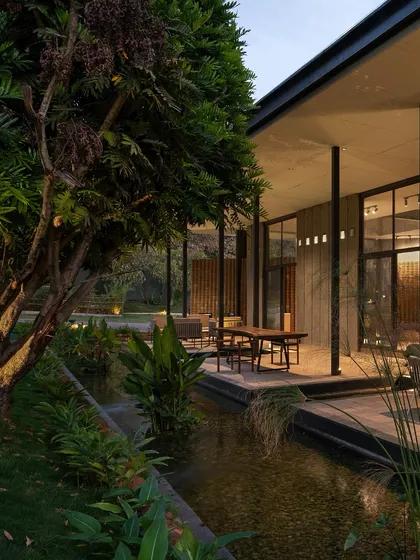 A tranquil water channel runs alongside the veranda of 'The Earth', reflecting the surrounding trees and the building's support columns, enhancing the connection between the structure and nature.