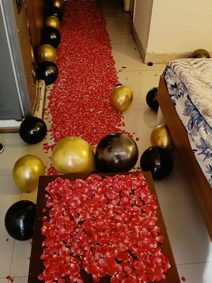 A romantic room setup featuring a long pathway of red rose petals leading to the bed, with black and gold balloons scattered around.