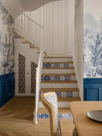This staircase, adjacent to the dining area, features custom-painted tile risers that echo the blue-and-white theme of the room. The deep indigo wainscoting adds a bold contrast to the natural wood floors.