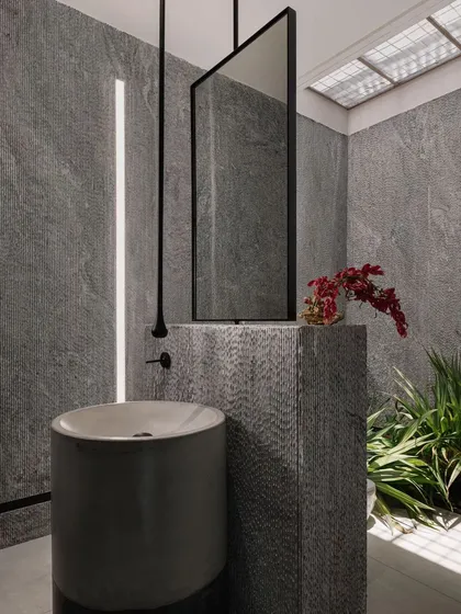This powder room is a dramatic space, clad in grey stone with a freestanding basin. A skylight and a vertical light slit illuminate the textures, creating a moody and sophisticated atmosphere.