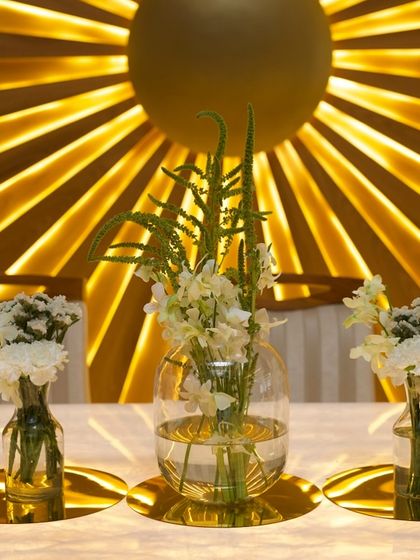 A simple yet stunning detail from a Roka ceremony. Minimalist floral arrangements in clear glass vases sit on a table in front of a dramatic, backlit golden backdrop, creating a modern and glamorous look.