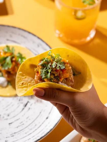 A hand holding a fish taco with mango salsa. This shot is interactive and highlights the fresh, seasonal ingredients.