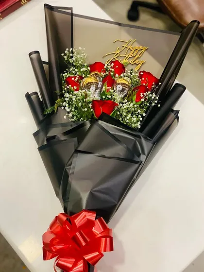 A detailed look at a birthday bouquet combining red roses and chocolates. The golden "Happy Birthday" topper adds a festive touch to the arrangement of flowers and Ferrero Rocher style chocolates.