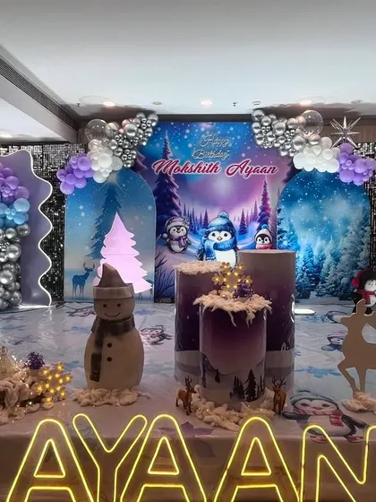 This is an example of a custom 'Winter Wonderland' theme I created for a birthday party. The setup includes a full stage backdrop with a winter forest scene, balloon arrangements in purple and silver, and themed props like a snowman and light-up name signage.