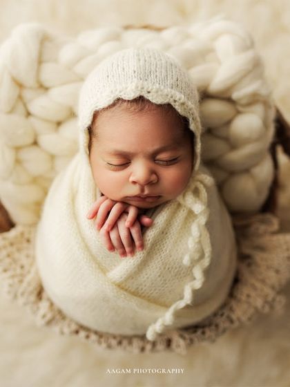 A classic newborn pose in a cozy basket. The baby is swaddled in white with a matching bonnet, their tiny hands resting gently under their chin. This is a perfect example of a timeless, angelic portrait.