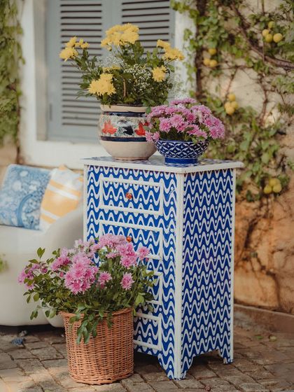 A detail from our sun-drenched bohemian Mehendi, featuring a hand-painted blue and white patterned side table, artisanal pots, and vibrant pink and yellow flowers.