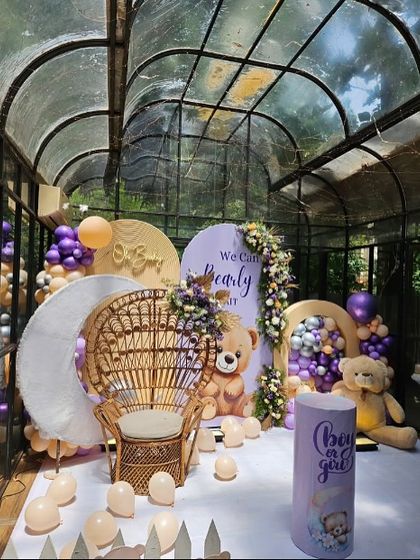 A stunning glasshouse venue decorated for a 'We Can Bearly Wait' baby shower. The setup includes a large wicker chair for the mom-to-be, multiple backdrops, and balloon decor in purple and pastel shades.