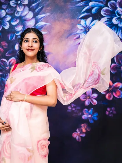 The mom-to-be looks ethereal in a delicate pink organza saree, with the fabric flowing around her against a painted floral background.