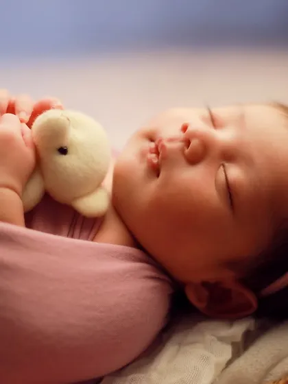 A close-up portrait showing the peaceful expression of a sleeping newborn clutching a small teddy bear. The soft lighting highlights her delicate features, from her lips to her eyelashes.
