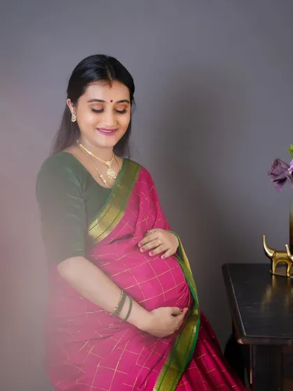 A soft, beautiful portrait of a mother-to-be in a pink silk saree, gently holding her baby bump with a loving expression.
