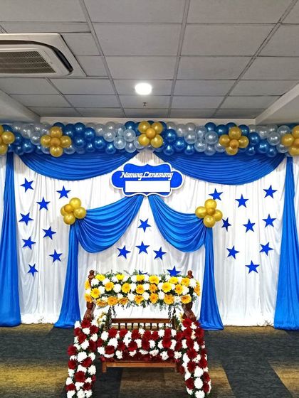 A naming ceremony stage decoration with a blue and white theme. The setup includes a flower-adorned cradle, draped curtains, and a balloon arch.