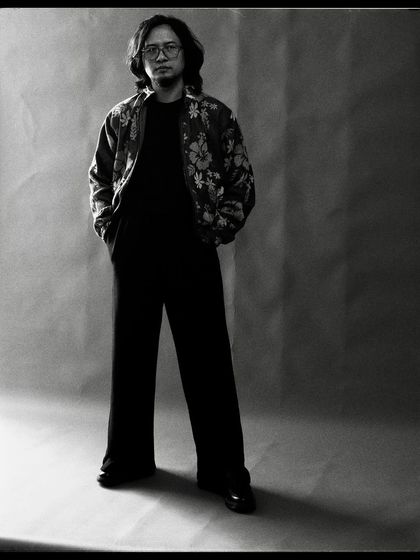 A full-length black and white portrait of Taba Chake. The simple studio background allows his quiet confidence to take center stage.