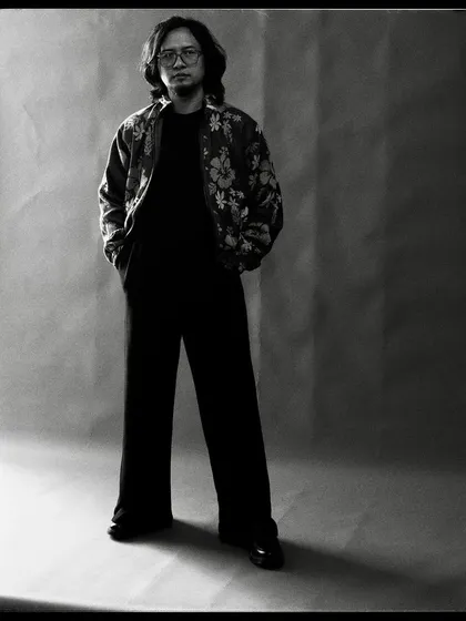 A full-length black and white portrait of Taba Chake. The simple studio background allows his quiet confidence to take center stage.