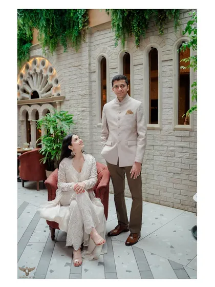 A classic, posed portrait in an elegant courtyard. The composition, with her seated and him standing, creates a regal and sophisticated image that highlights their coordinated outfits and the stunning architectural details.
