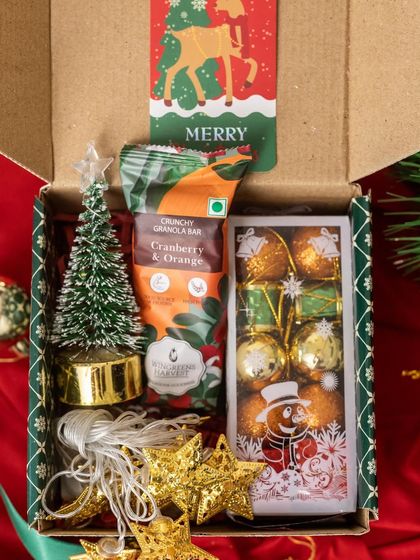 A delightful Christmas hamper filled with light and sweetness. This box contains a mini Christmas tree, a string of fairy lights, a crunchy granola bar, and festive chocolates.