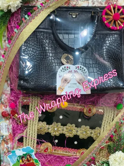 A Teej trousseau packing that includes a stylish black handbag and a decorated saree, all arranged on a platter with pink shredded paper.
