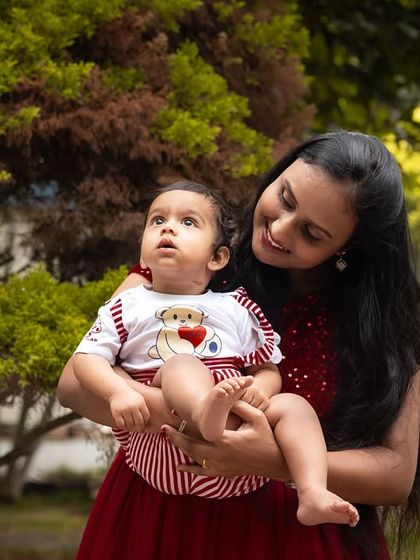 A mother and her baby share a moment during an outdoor photoshoot. The natural green background provides a beautiful contrast to their red outfits.