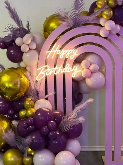 A trendy and glamorous birthday setup featuring a pink rainbow arch prop. The decor is elevated with a "Happy Birthday" neon sign and a rich balloon arrangement in shades of purple, pink, and gold.