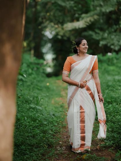 A beautiful portrait of the bride in her kasavu saree with an orange blouse, smiling amidst the greenery.