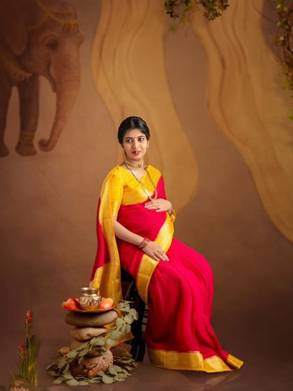 A serene portrait of a mom-to-be in a vibrant red and yellow saree, seated against a painterly backdrop featuring an elephant.