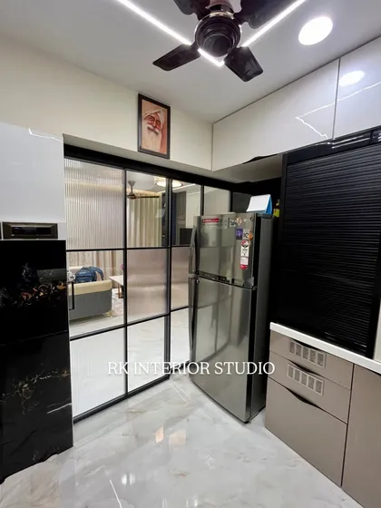 A view of a modern kitchen with a glass partition separating it from the living area. The kitchen features a mix of white and beige cabinets, with a black roller shutter for appliances.