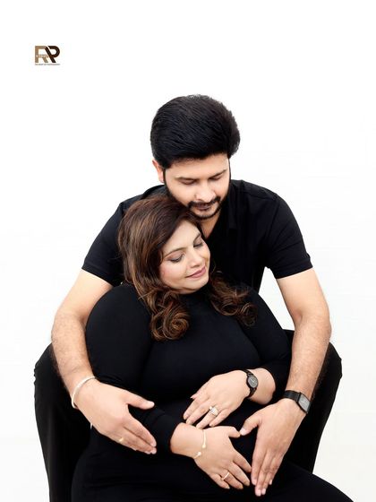 A tender moment captured in a modern maternity shoot. The husband-to-be embraces his wife from behind, both dressed in black for a simple and elegant portrait.