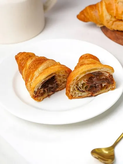 A delicious Nutella-filled croissant, cut in half to show the generous filling, placed on a white plate with a golden spoon. This shot is perfect for a bakery or cafe's dessert menu, highlighting the indulgent treat.