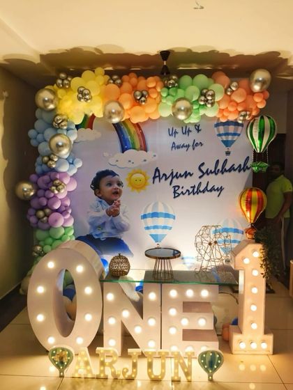 A dreamy 'Up, Up and Away' hot air balloon theme for a first birthday. This setup features a custom backdrop, large marquee letters, and a pastel rainbow balloon garland.