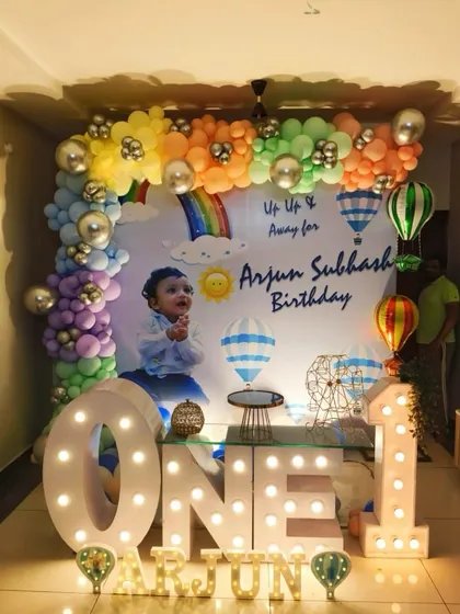 A dreamy 'Up, Up and Away' hot air balloon theme for a first birthday. This setup features a custom backdrop, large marquee letters, and a pastel rainbow balloon garland.