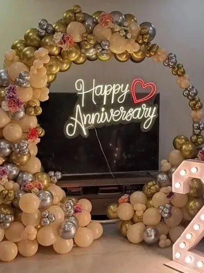 A 25th-anniversary setup with a balloon arch in beige, gold, and silver, framing a 'Happy Anniversary' neon sign.