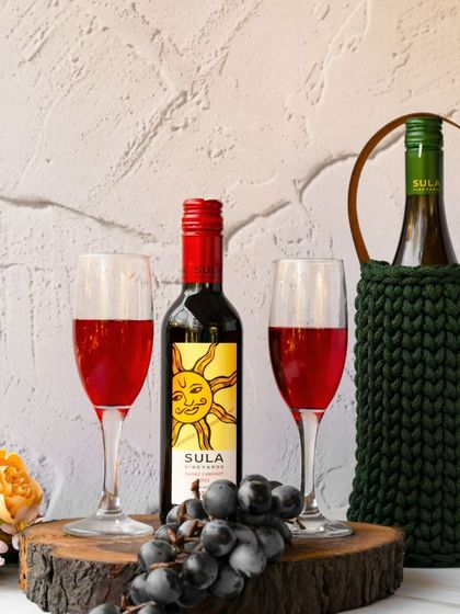 A romantic wine setting featuring two glasses of red wine, grapes, and two bottles of Sula. One bottle is nestled in a dark green hand-crocheted wine bag, adding a touch of handmade charm.