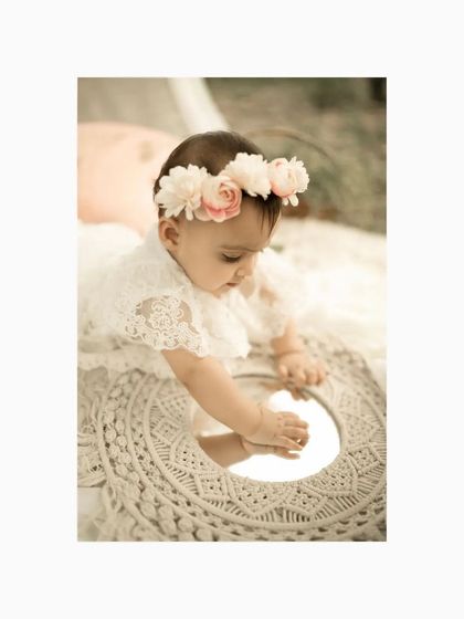 A close-up shot of the baby girl, wearing a floral crown and looking at her reflection in a small mirror. This captures a sweet moment of self-discovery during her outdoor milestone session.