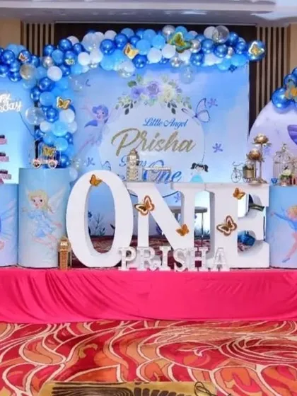 A classic first birthday celebration with a "Little Angel" theme. The decor features large illuminated "ONE" letters, a blue and white color scheme, and butterfly accents on the backdrop and pedestals.