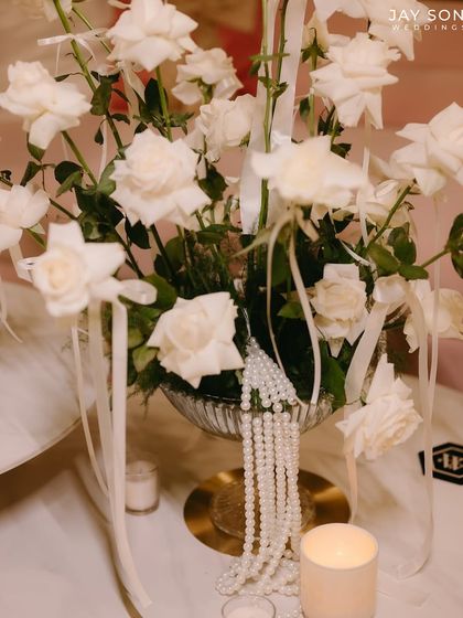 A centerpiece that embodies the "Big Bow Affair" theme. A classic arrangement of white roses is elevated with delicate pearl strings and small white ribbons tied to each stem.
