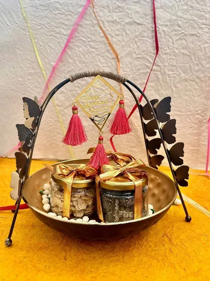 A modern and chic Sankranti hamper in a butterfly-handled basket. This gift contains jars of til-gud and other festive sweets, perfect for celebrating the harvest festival in style.